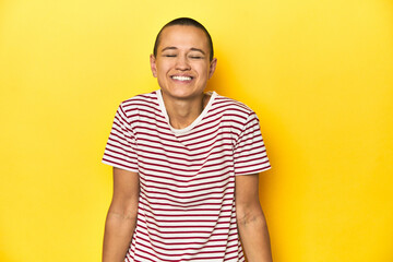 Shaved head woman in red striped tee, yellow backdrop laughs and closes eyes, feels relaxed and happy.