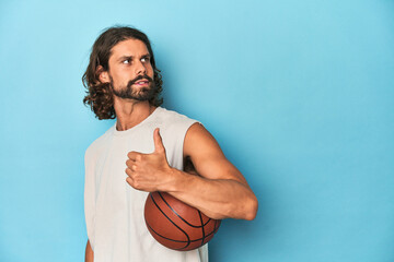 Bearded man with basketball in blue studio points with thumb finger away, laughing and carefree.