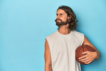Bearded man with basketball in blue studio looks aside smiling, cheerful and pleasant.