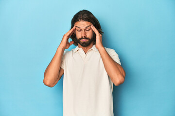 Long-haired man in a white polo, blue studio touching temples and having headache.