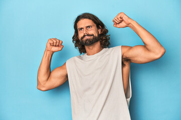 Naklejka premium Bearded man in a tank top, blue backdrop raising fist after a victory, winner concept.