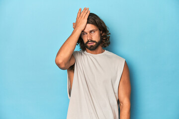 Bearded man in a tank top, blue backdrop forgetting something, slapping forehead with palm and closing eyes.