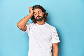 Bearded man in a white shirt, blue backdrop tired and very sleepy keeping hand on head.