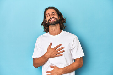 Bearded man in a white shirt, blue backdrop laughs happily and has fun keeping hands on stomach.