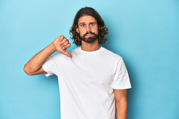 Bearded man in a white shirt, blue backdrop showing a dislike gesture, thumbs down. Disagreement concept.