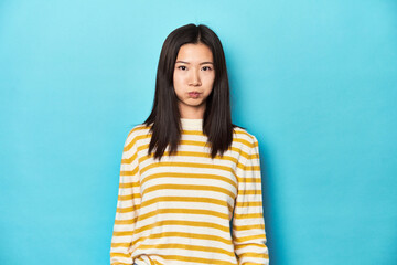 Asian woman in striped yellow sweater, blows cheeks, has tired expression. Facial expression...