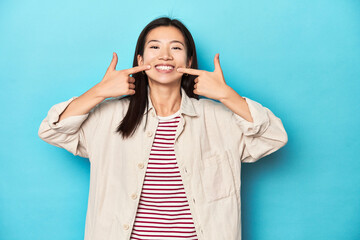Asian woman in layered shirt and striped t-shirt, smiles, pointing fingers at mouth.