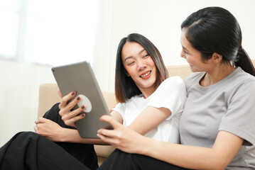 Affection LGBT concept. Happy diverse beautiful asian female lesbian couple talking and using tablet in living room in slow motion shot. Spending quality time, joy, technology and love.