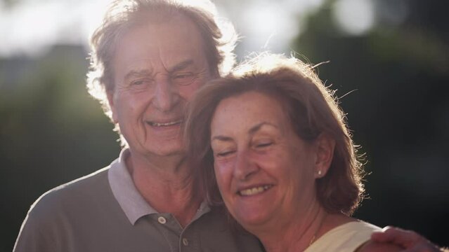 A Happy Married Older Couple Looking At Camera At Park With Sunlight Flare Backlight. Close Up Of Senior Faces Of 70s Husband And Wife