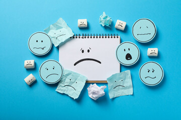 Notepad and papers with sad emoji on blue background, top view
