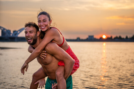 Happy young couple in love having fun relaxing on beach near city. Caucasian girlfriend piggybacking on boyfriend enjoying holidays together. Selective focus