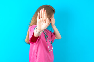 Young caucasian doctor woman wearing pink medical uniform covers eyes with palm and doing stop...