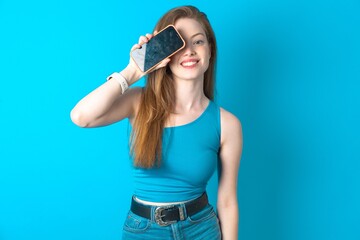 Young beautiful girl wearing blue T-shirt holding modern smartphone covering one eye while smiling