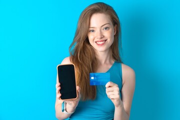 Photo of adorable Young beautiful girl wearing blue T-shirt holding credit card and Smartphone. Reserved for online purchases