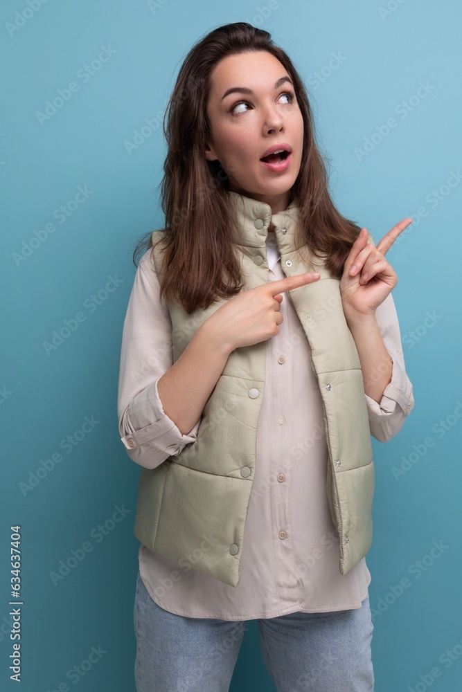 Sticker young brunette woman in a shirt and vest points a finger for ideas and inspiration on a background with copy space