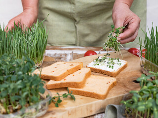 A woman prepares a sandwich with milk paste and microgreens. Nearby there are many boxes with a variety of microgreens. Healthy food concept.