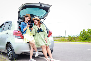 Two Asian women stand behind the car smiling happily looking at the photo together. Asian tourists. Travel concept, transport. copy space