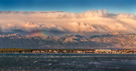 Vrsi, Croatia - Panoramic view of Vrsi municipality in Zadar county with Velebit mountains and Paklenica national park covered with warm golden colored clouds and rainbow in background at sunset