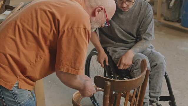 Medium shot of young Caucasian wheelchaired carpenter restoring old wooden furniture with workmate in carpentry workshop