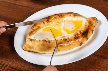 Man eating adjar Khachapuri. Georgian national pie khachapuri with egg and cheese in the white plate  on wooden background.