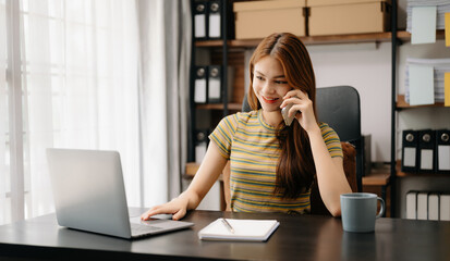 Business asian woman Talking on the phone and using a laptop with a smile while sitting at home office.