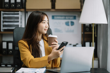 Asian businesswoman sits in a office working on smartphone and enjoys a coffee.