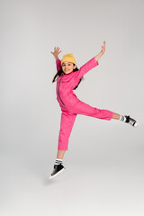 happy girl in beanie hat and stylish pink outfit jumping with outstretched hands on grey, have fun