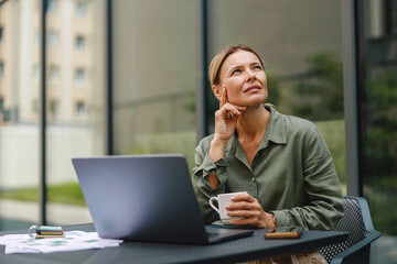 Focused woman sales manager working on laptop and drinking coffee in terrace and looking at side