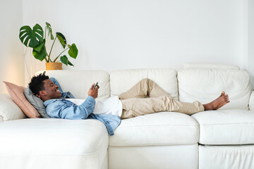Smiling black man using smartphone and lying on cozy sofa