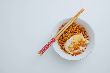 Flat lay or top view shot of a portion of stir-fried Indonesian instant noodles with egg on a white bowl with a pair of wooden chopsticks on white table