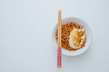 Flat lay or top view shot of a portion of stir-fried Indonesian instant noodles with egg on a white bowl with a pair of wooden chopsticks on white table