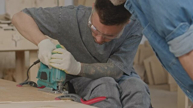 Medium shot of concentrated wheelchaired woodworker using jigsaw while cutting wooden plank, making furniture with workmate in carpentry studio