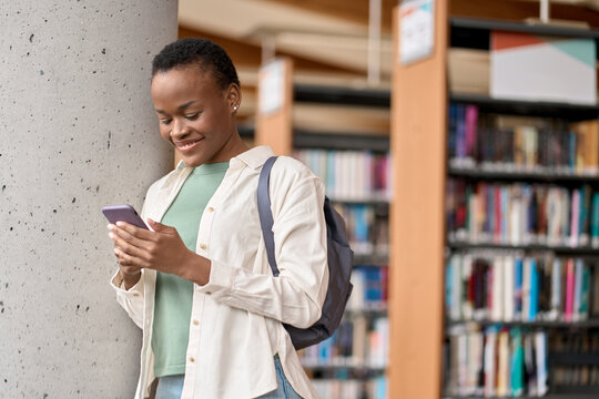 Happy African Black Teen Girl College Generation Z Student With Backpack Looking At Smartphone Using Mobile Cell Phone Modern Tech Device Standing In University Campus. Copy Space.