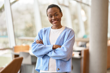 Happy cute African teenage girl, smiling confident short-haired cute Black ethnic college student standing arms crossed looking at camera in modern foreign university campus studying abroad. Portrait.