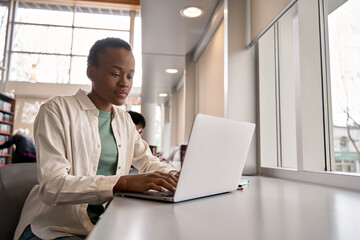 Busy short-haired African girl student using laptop technology sitting in university campus library. Black teenager elearning looking at computer doing coursework hybrid learning online research.