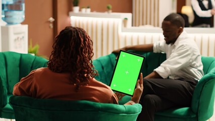 Happy guests in hotel lounge holding chroma key green screen mock up tablet while enjoying cup of coffee. African american couple waiting to be checked in, sitting on cozy sofa