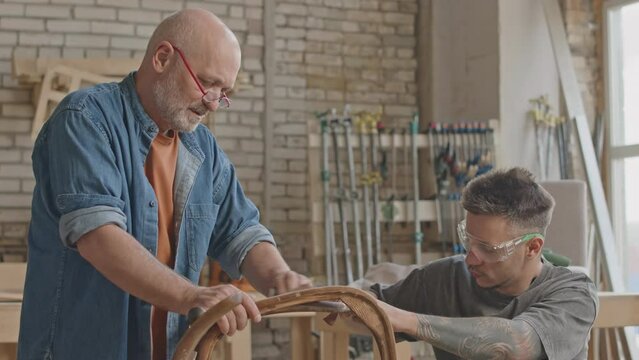 Medium shot of two professional carpenters sanding wooden surface and legs of handmade chair by hand, working together in carpentry workshop