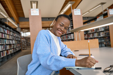 Happy smiling African Black girl college student holding pencil in hand writing in paper notebook, taking notes, studying, learning in modern university library looking at camera. Portrait.