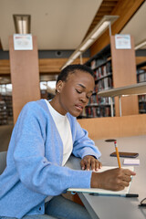 African Black girl college student holding pencil in hand writing in paper notebook, taking notes, studying, learning in modern university library doing homework. Vertical shot.