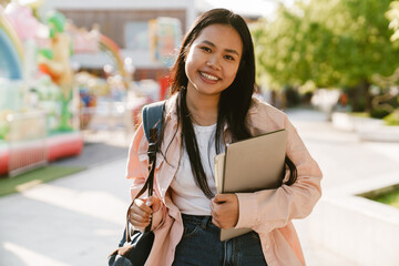 Asian woman with backpack and laptop smiling at camera while standing outdoors