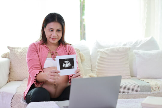 Happy smiling young asian pregnant woman resting and sitting on sofa in living room while showing ultrasound image with computer. Expectant mother preparing and waiting for baby birth during pregnancy