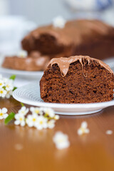 Homemade chocolate cake with chocolate glaze on white plate on laid table with bouquet of spring blossom apple tree branches