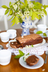 Homemade chocolate cake with chocolate glaze on white plate on laid table with bouquet of spring blossom apple tree branches