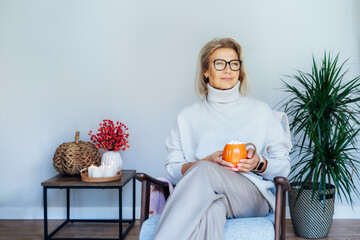 Middle aged woman relaxing with pumpkin shaped cup of hot drink in scandy style hygge interior home...