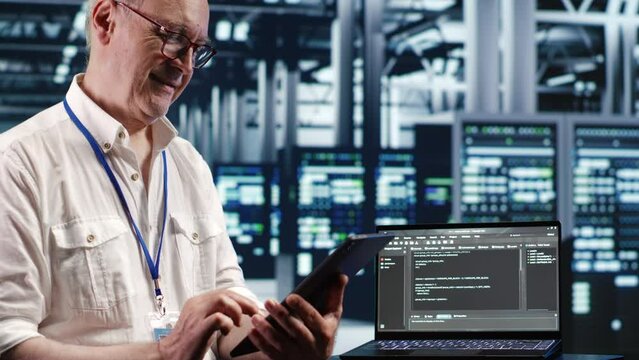 Elderly worker in data center, solving tasks on tablet while running scripts on laptop terminal. Certified engineer amidst servers, accessing databases and manipulating lines of code
