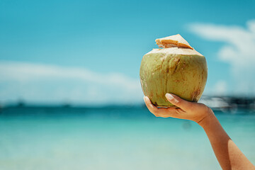 A woman holds a coconut on the background of the sea. Coconut cocktail on the beach. Azure sea - obrazy, fototapety, plakaty