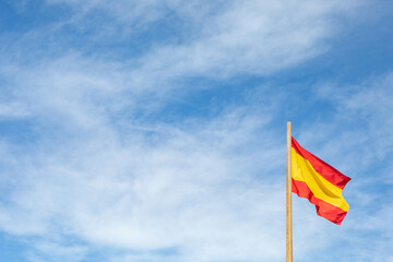 spanish flag on blue sky with white clouds and wooden mast.