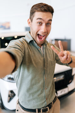 Vertical POV picture of cheerful buyer male taking selfie with victory sign on smartphone from auto dealership after bought new car. Smiling young man choosing new vehicle in showroom and making photo