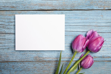 Invitation card mockup, white blank greeting card with purple tulip flowers on blue wooden background, flat lay. Floral greeting card mockup for a rustic touch