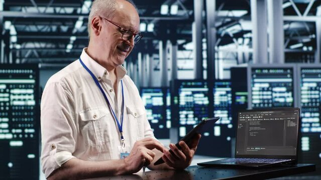Old system administrator in data center, solving tasks on tablet while running scripts on laptop terminal. Qualified employee amidst servers, accessing databases and manipulating lines of code